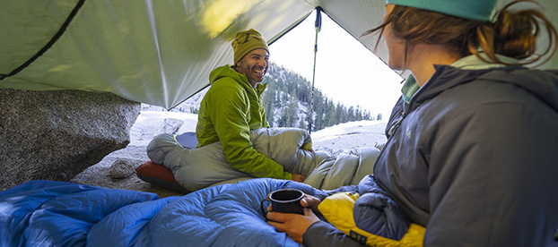 Un homme et une femme dans une tente, sous leurs sacs de couchage, avec une tasse à la main